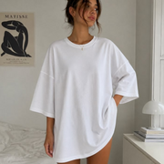 A woman wearing an oversized white short-sleeve t-shirt dress with side pockets, standing in a bright bedroom setting.